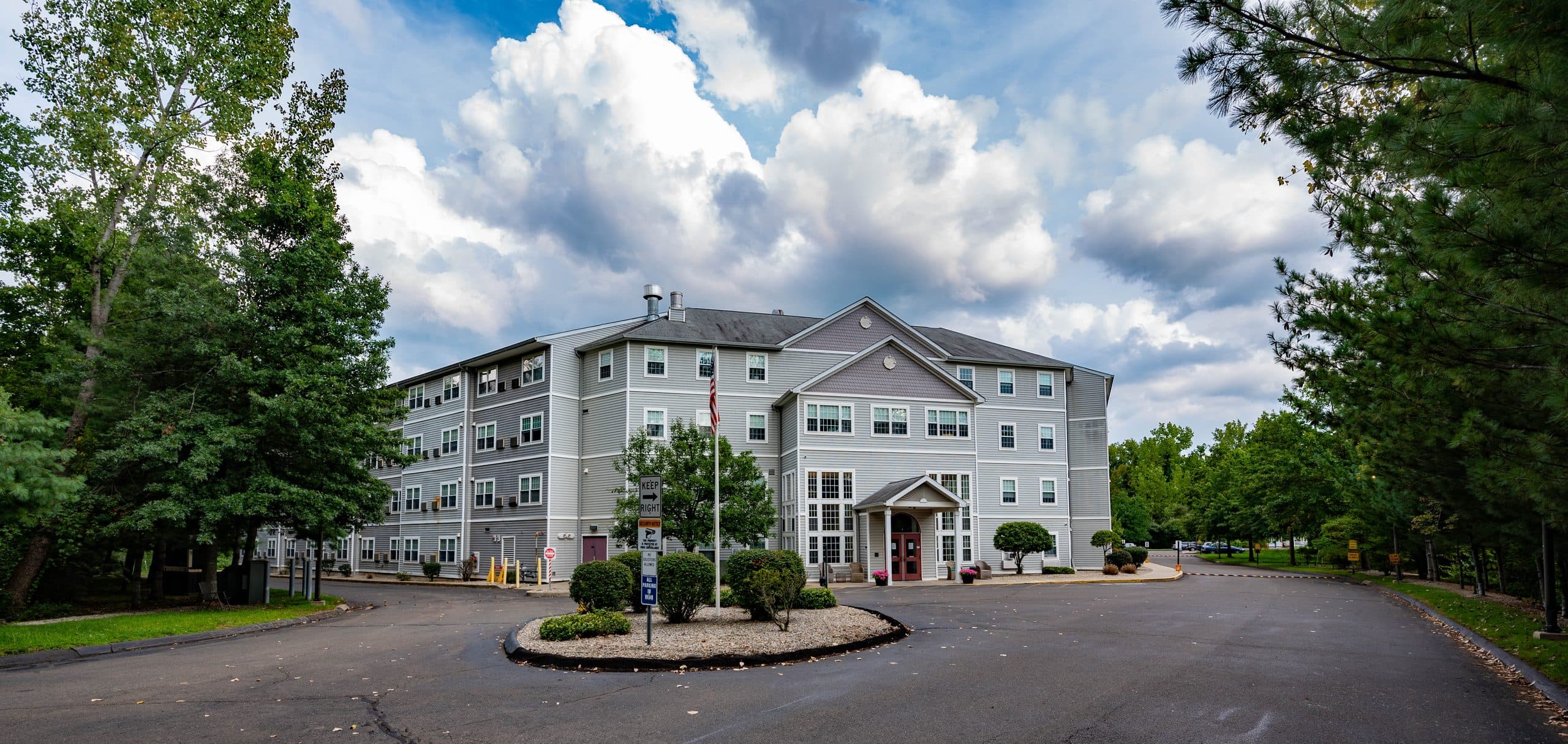 Carmen Romano Apartments — a four-storey clapboard building behind its circular drive and flagpole, framed by mature trees