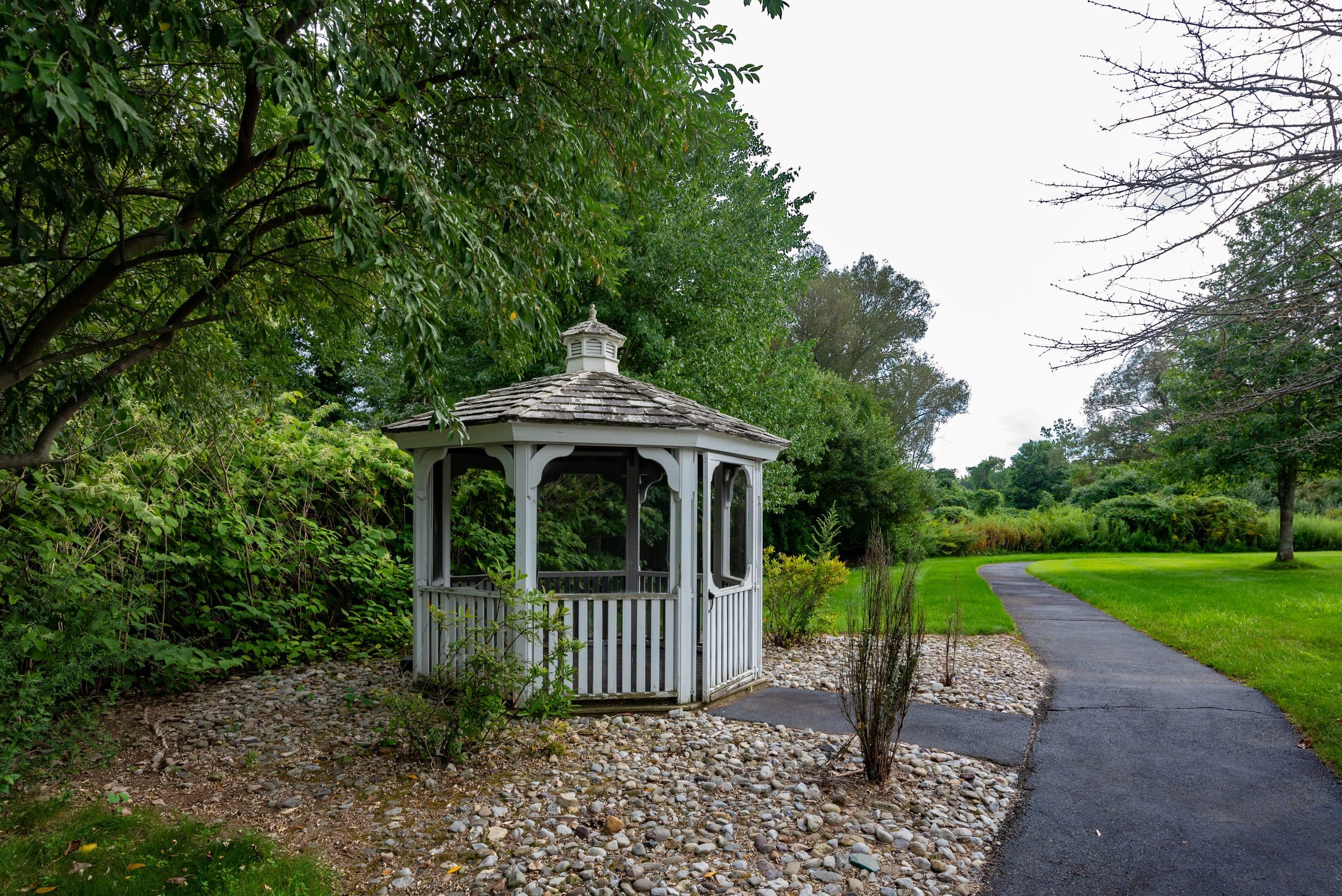 A white garden gazebo beside a walking path, surrounded by trees and plantings